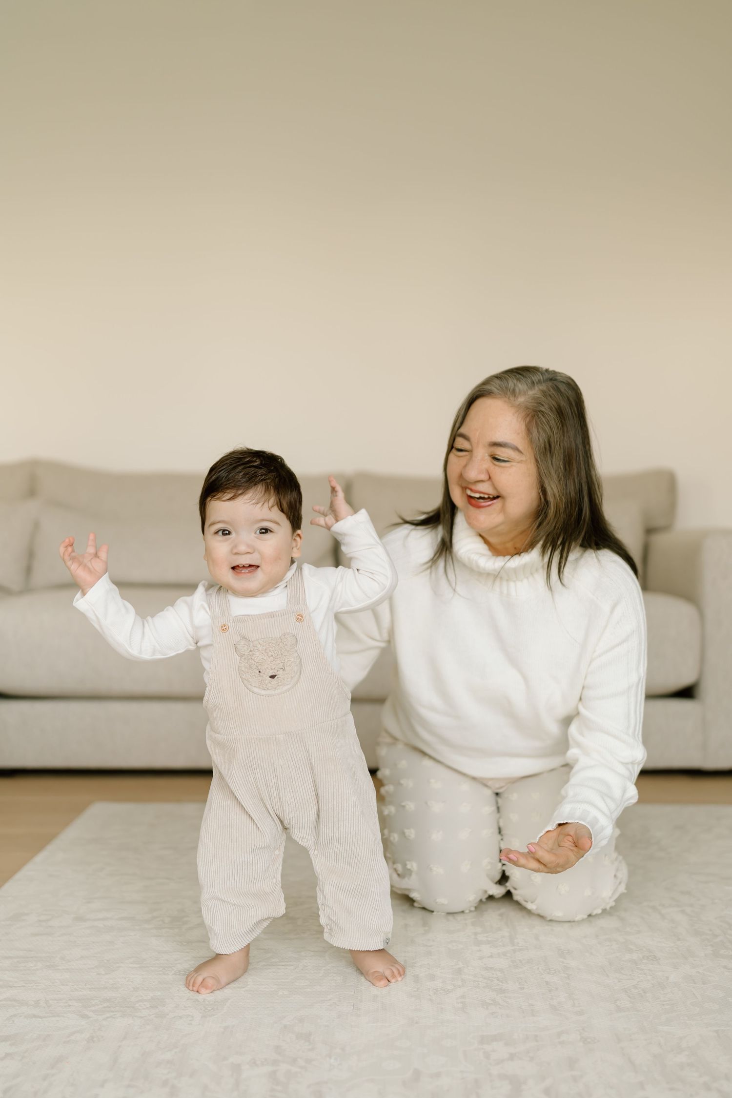 Two babies in white outfits laughing and playing together on a light colored carpet.