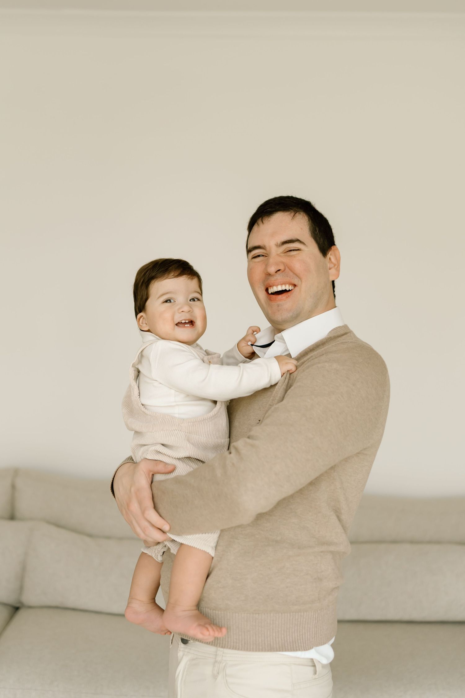 A heartwarming sequence of candid moments between a parent and baby wearing white, sharing joyful laughter on a beige couch.