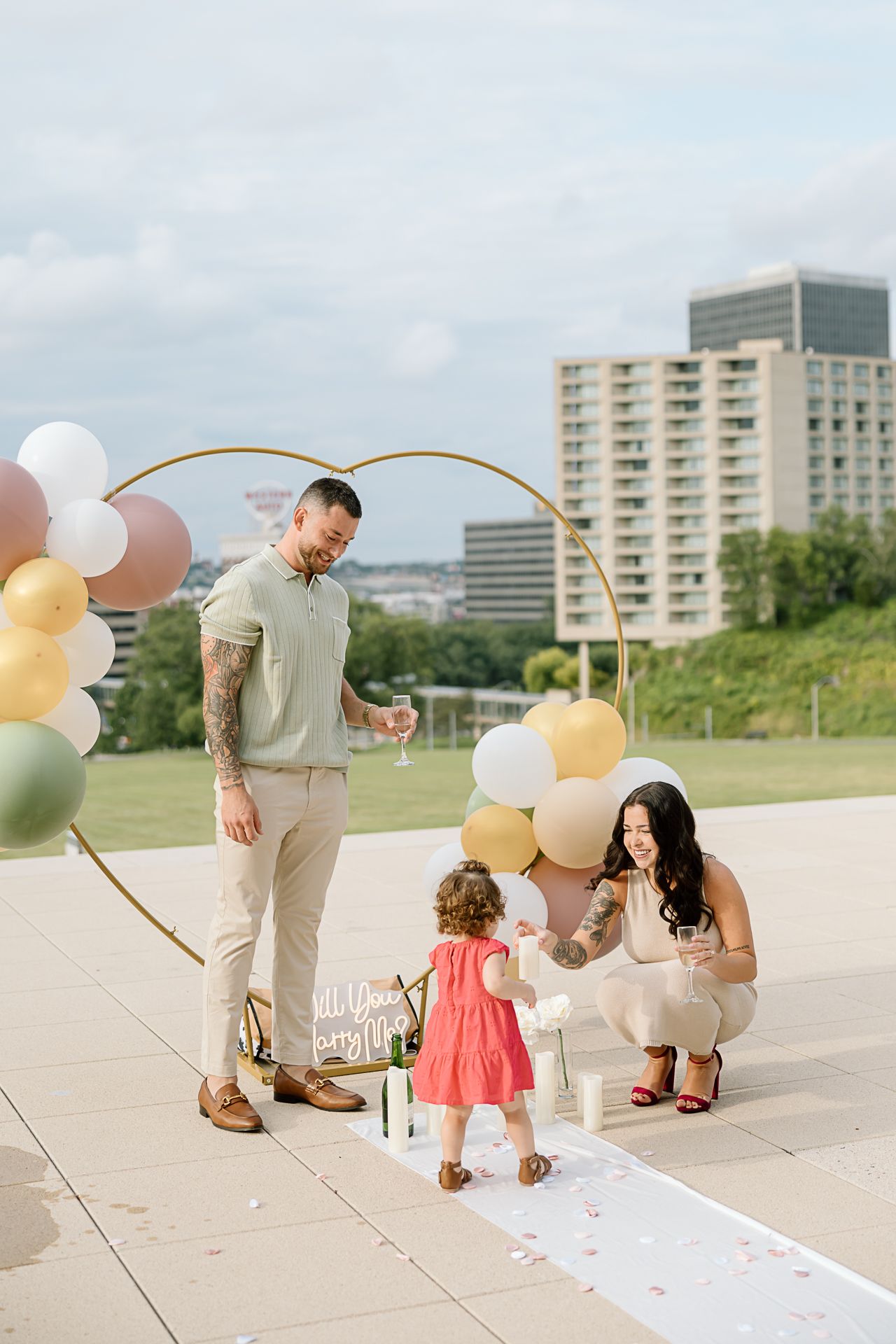 A couple celebrates with balloons and a sign on an outdoor plaza with city buildings in the background.
