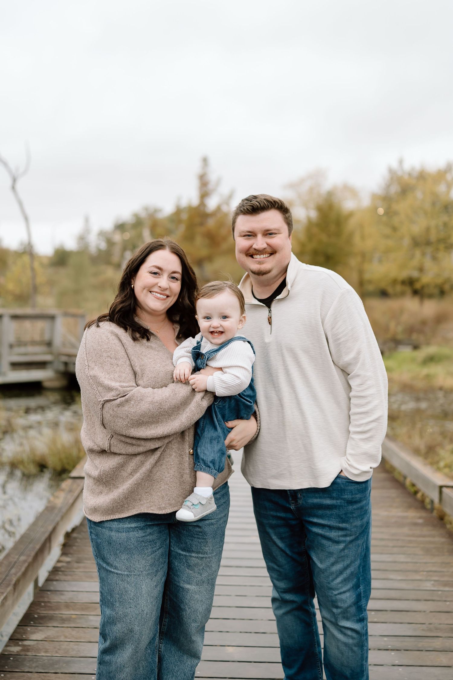 A family wearing coordinating beige sweaters and jeans poses together on a wooden boardwalk during an autumn photoshoot.