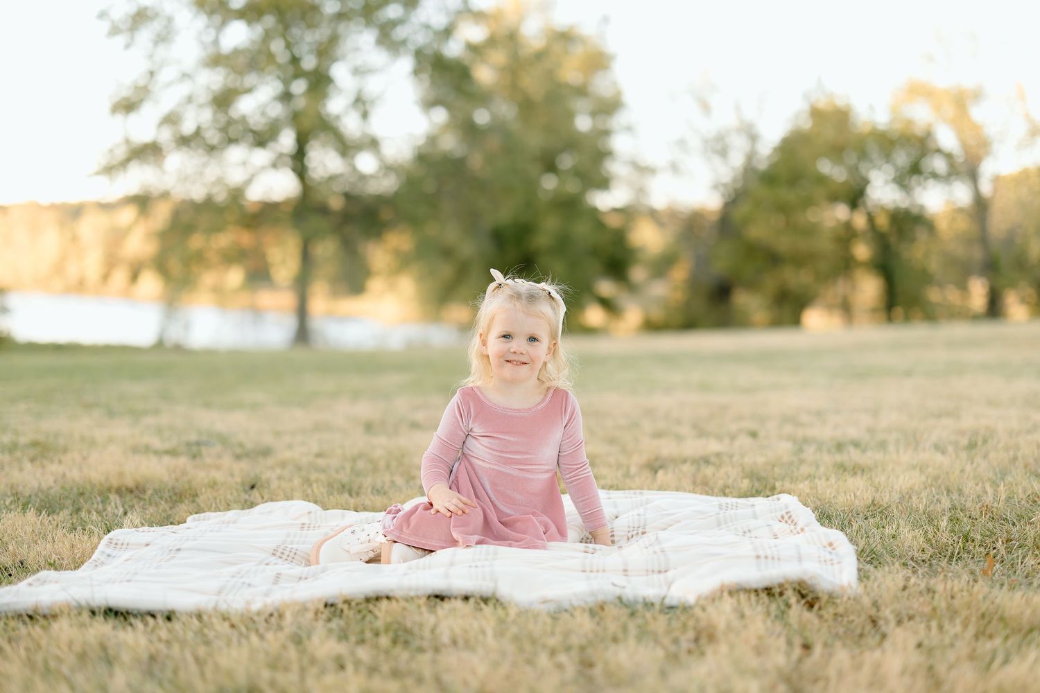 A child in a pink dress sits on a white blanket in a sunlit grassy field during golden hour.