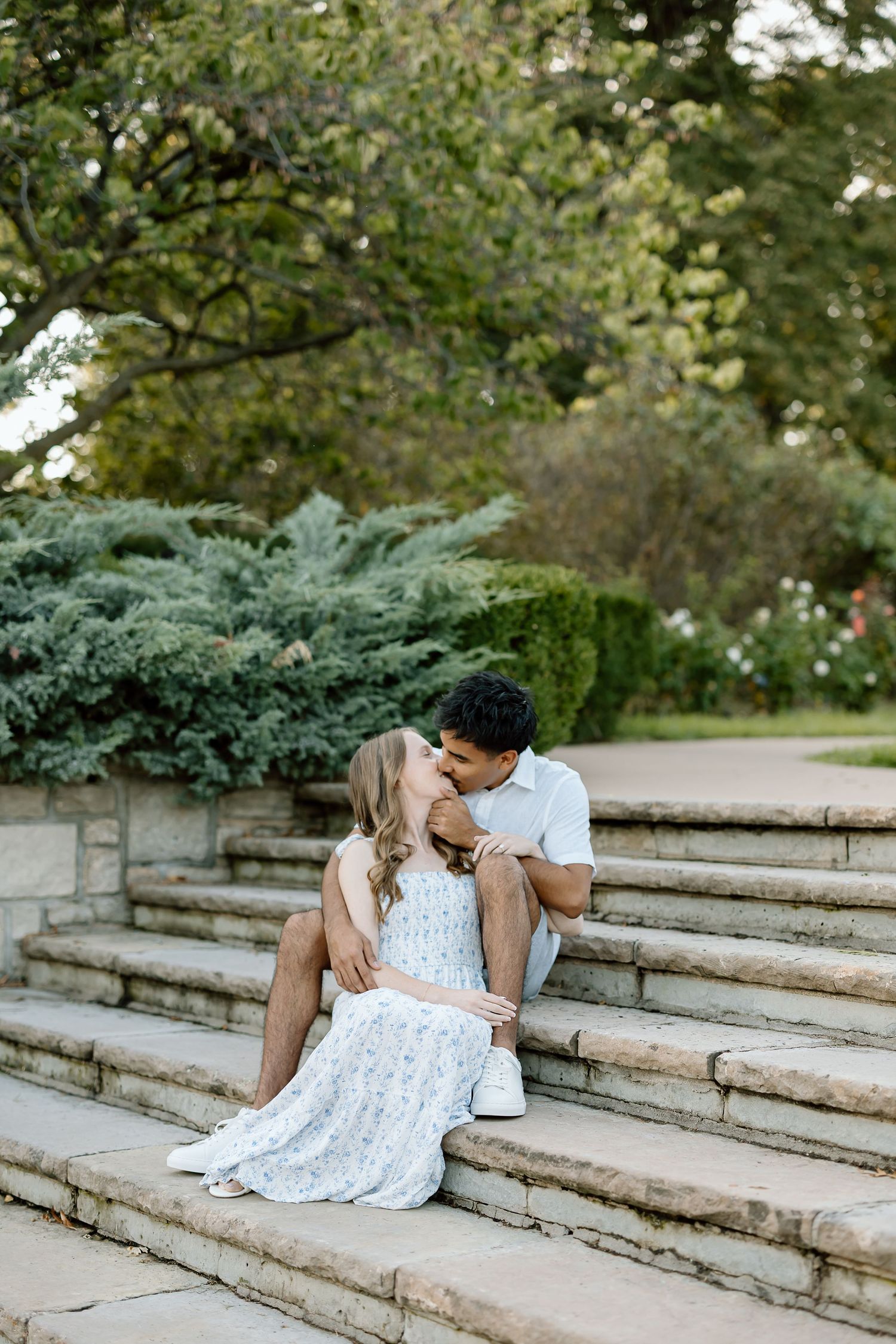 A couple sits intimately together on stone steps surrounded by lush greenery in a romantic outdoor setting.
