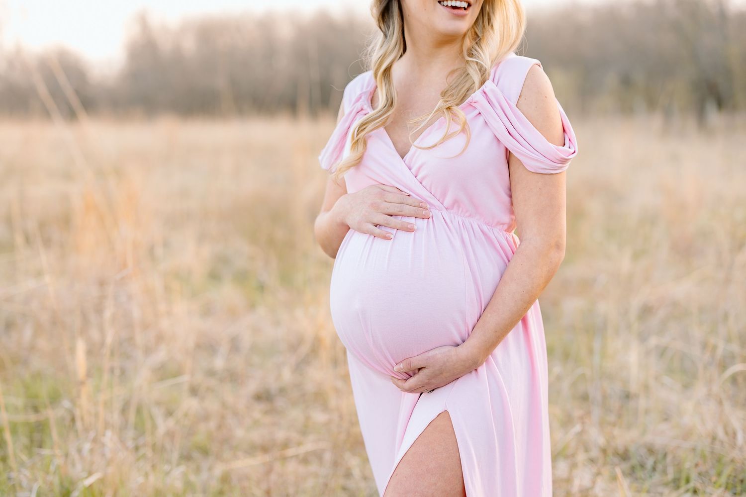A person in a pink dress cradles their pregnant belly while standing in a sunlit field at golden hour.