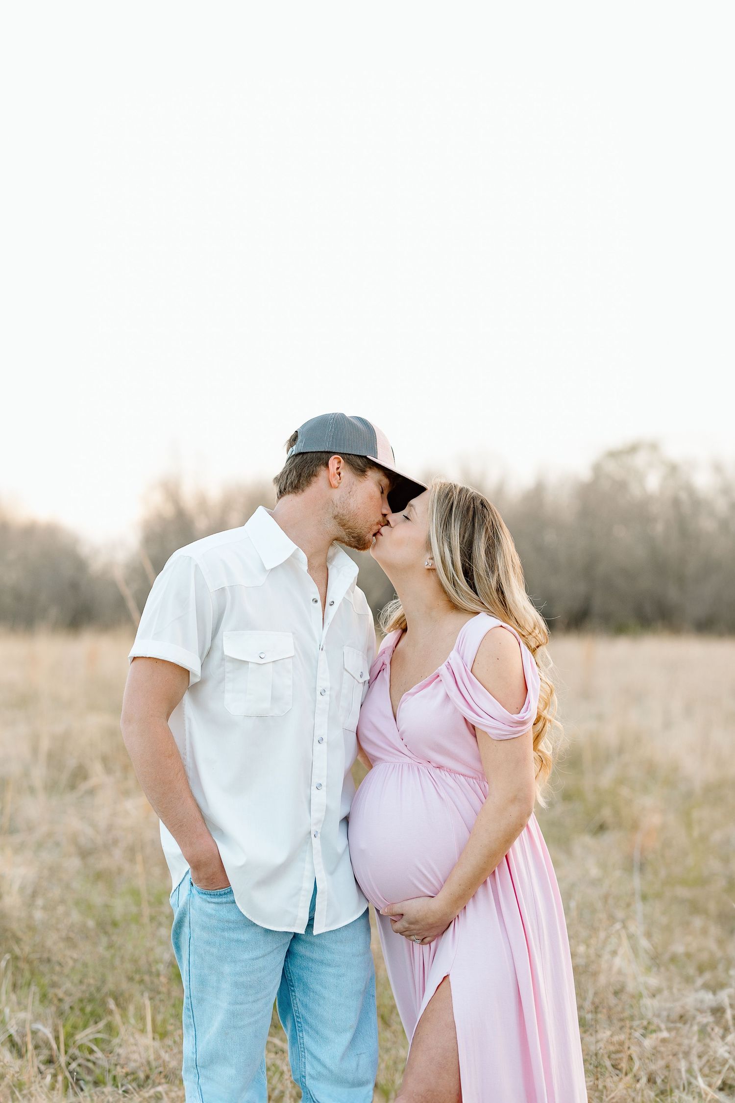 Romantic couple in a field at sunset with woman wearing pink maternity dress and man in white shirt and baseball cap.