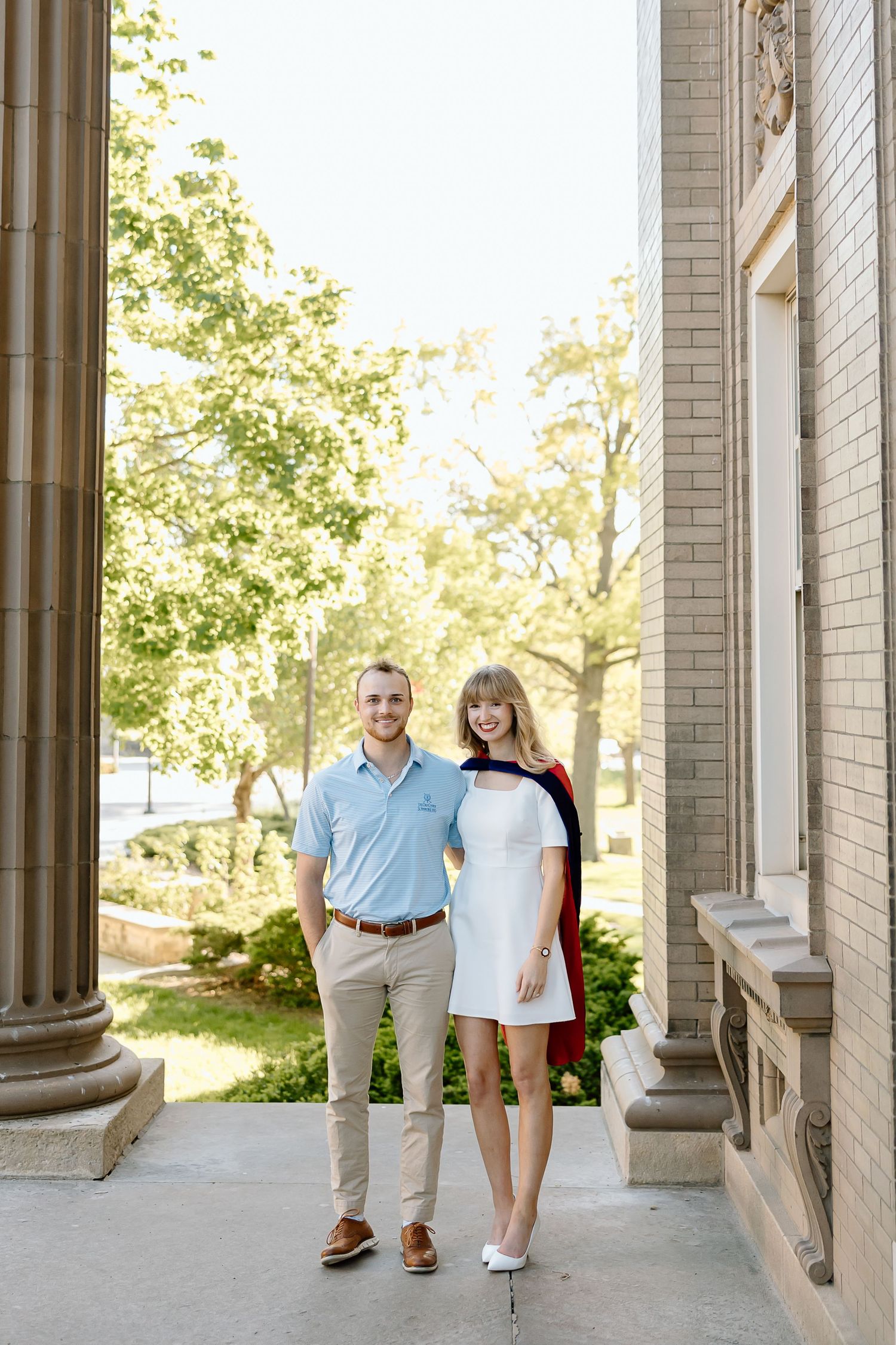 A couple poses for engagement photos under a columned walkway on a sunny day at a university campus.