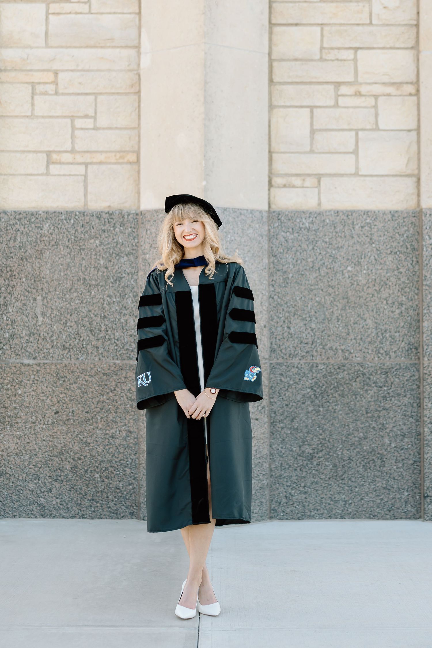 Graduate in black academic regalia poses in front of stone wall wearing white ballet flats.