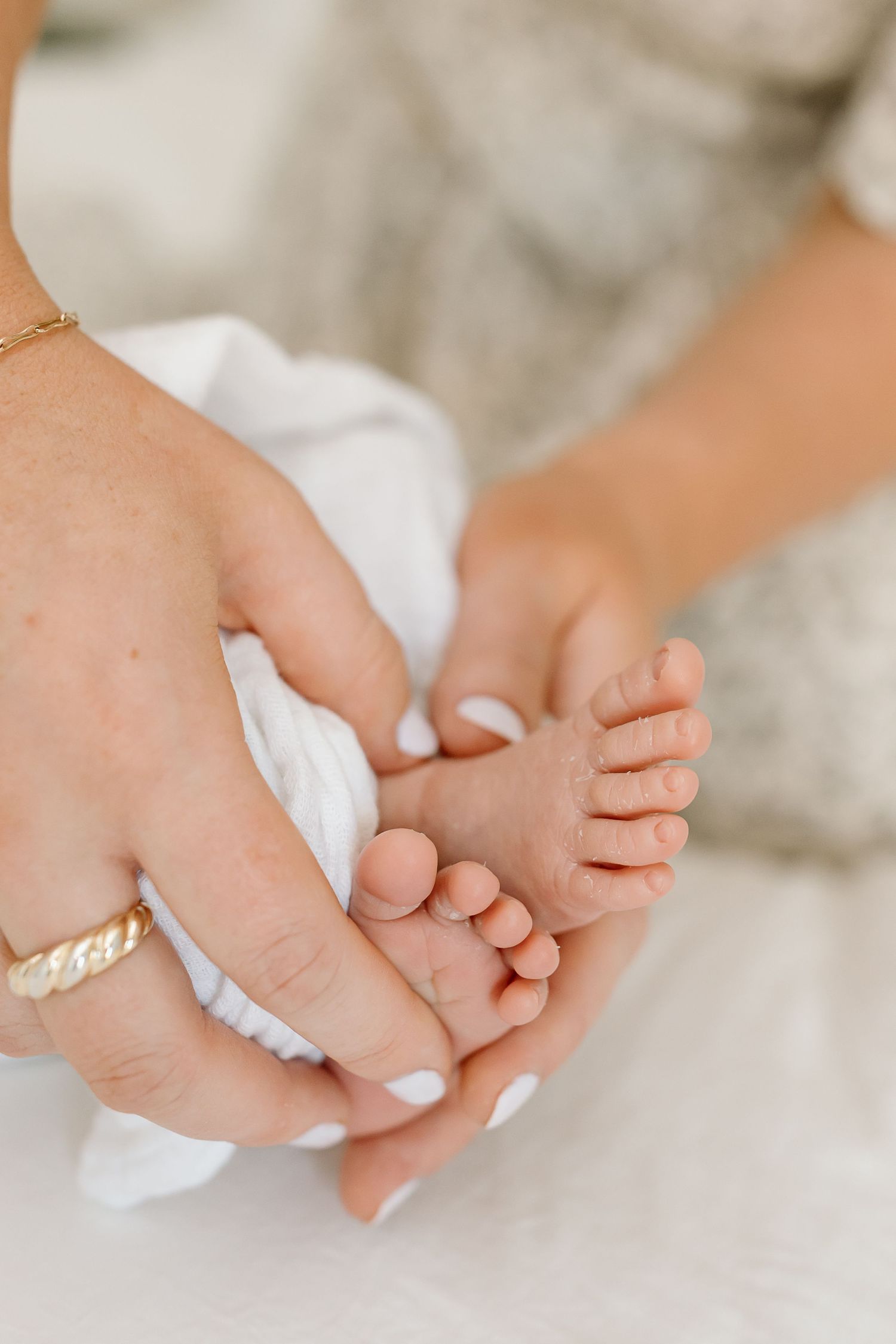 Gentle hands with white manicured nails holding tiny newborn baby feet wrapped in soft white fabric.
