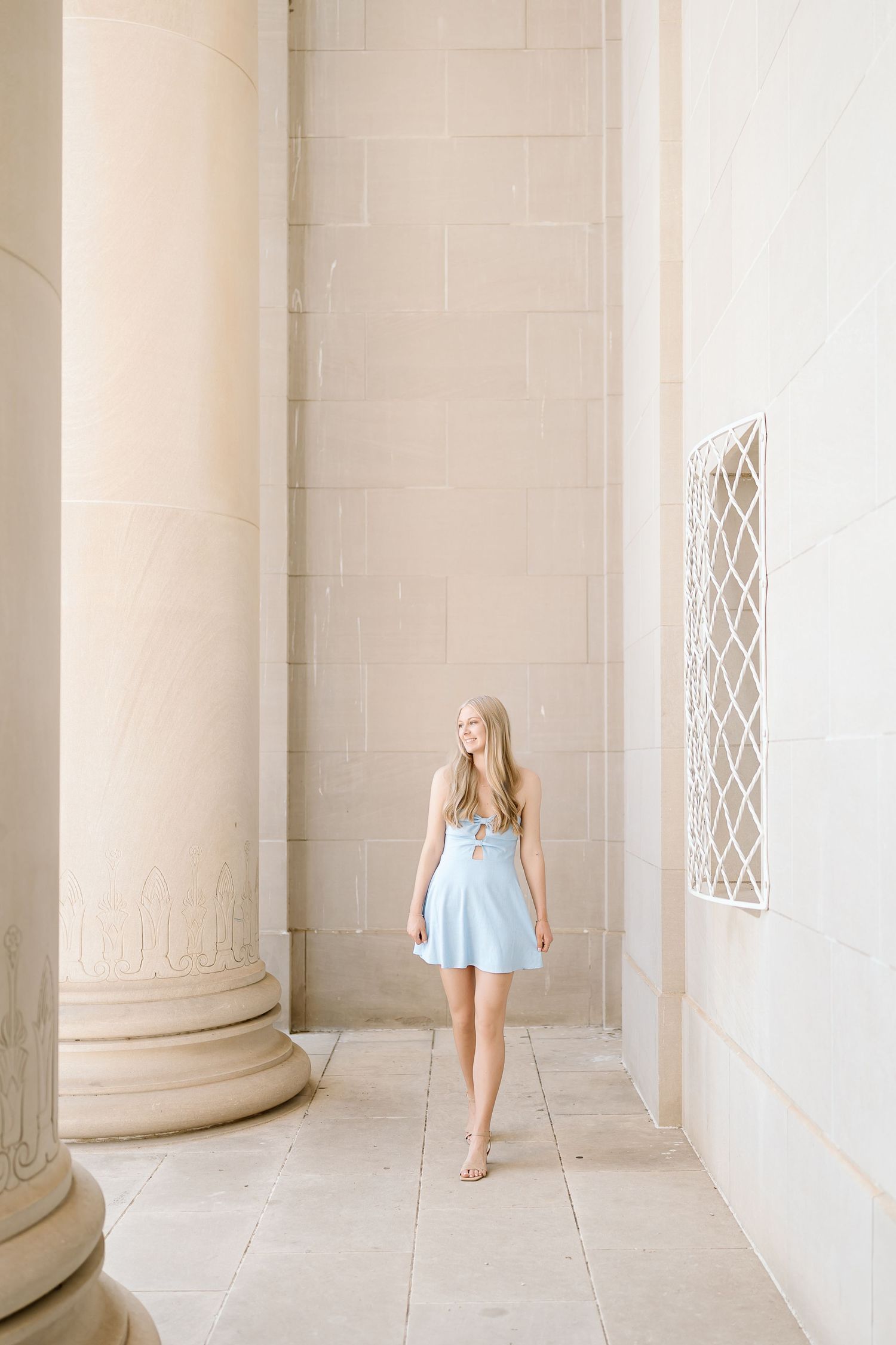 A person in a light blue dress walks through a grand colonnade with white stone columns and arches.