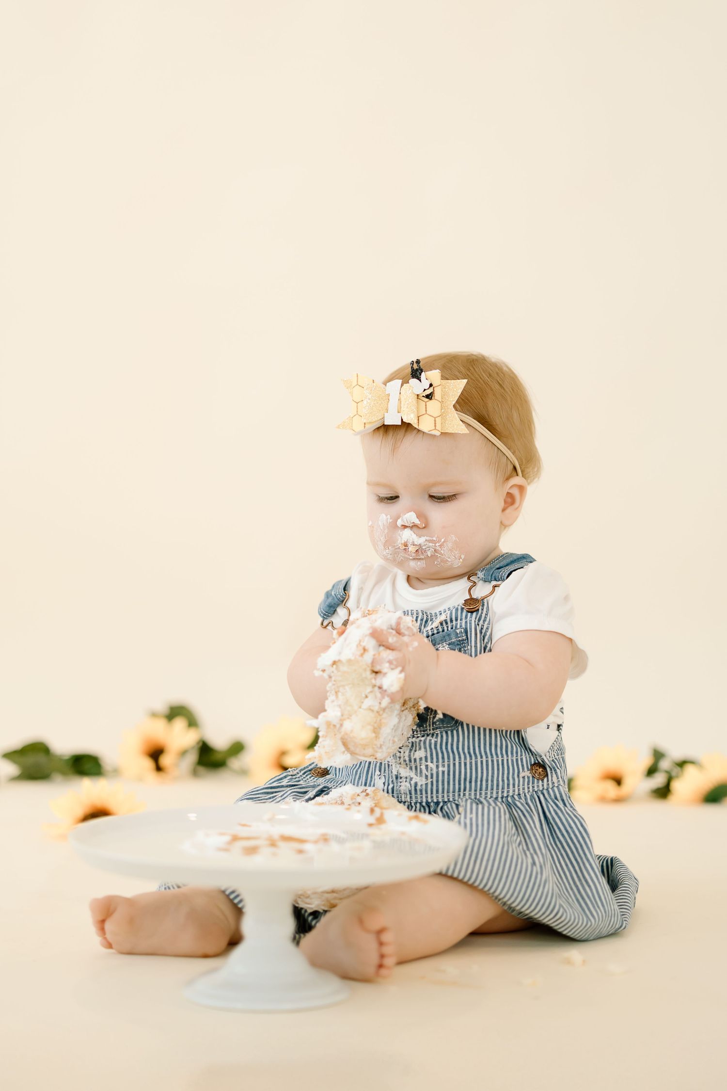 A baby in blue and white stripes enjoys a cake smash photo session with a white cake on a cream background.