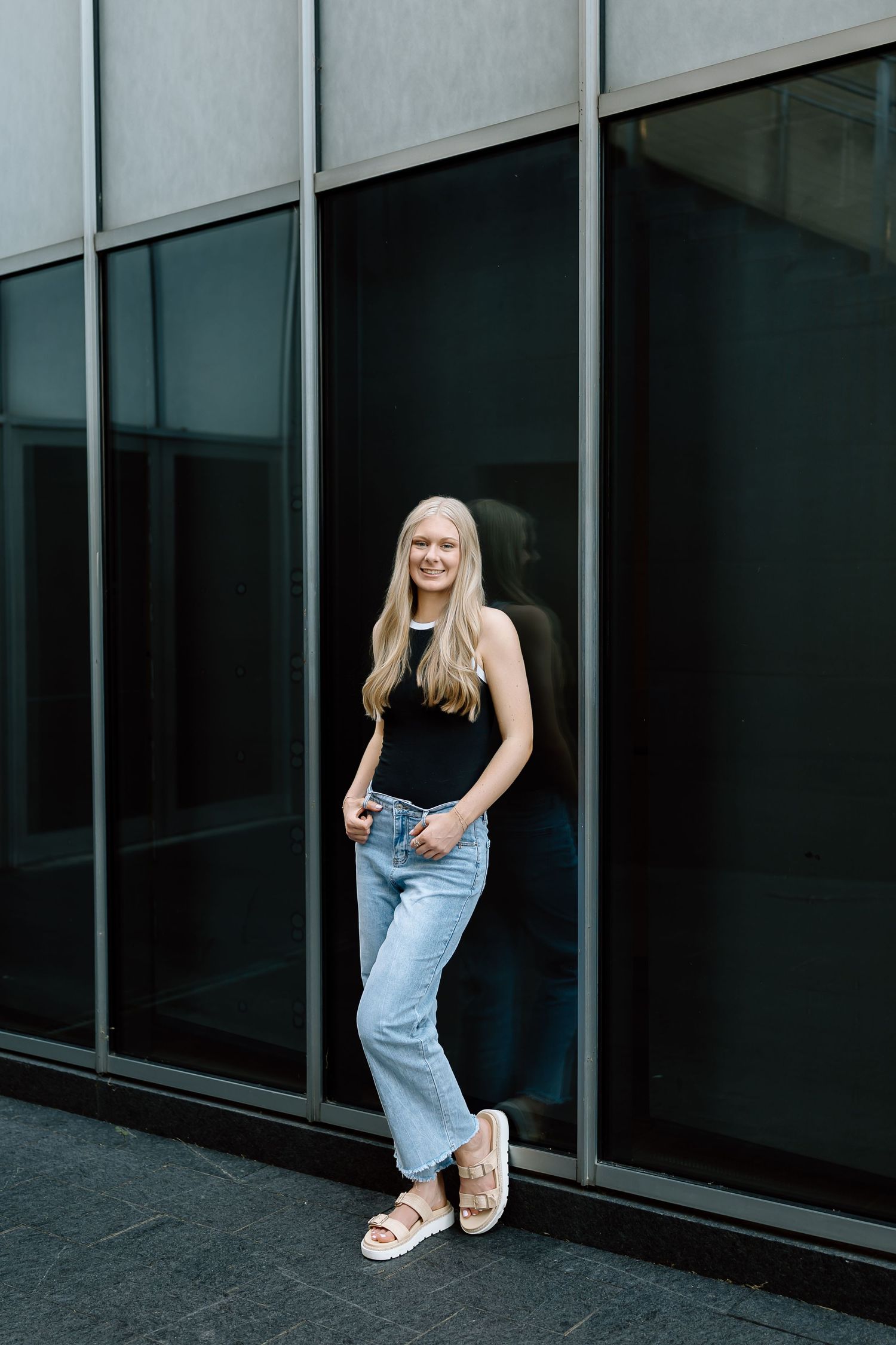 Woman in denim overalls and white sneakers poses playfully against modern glass building exterior.