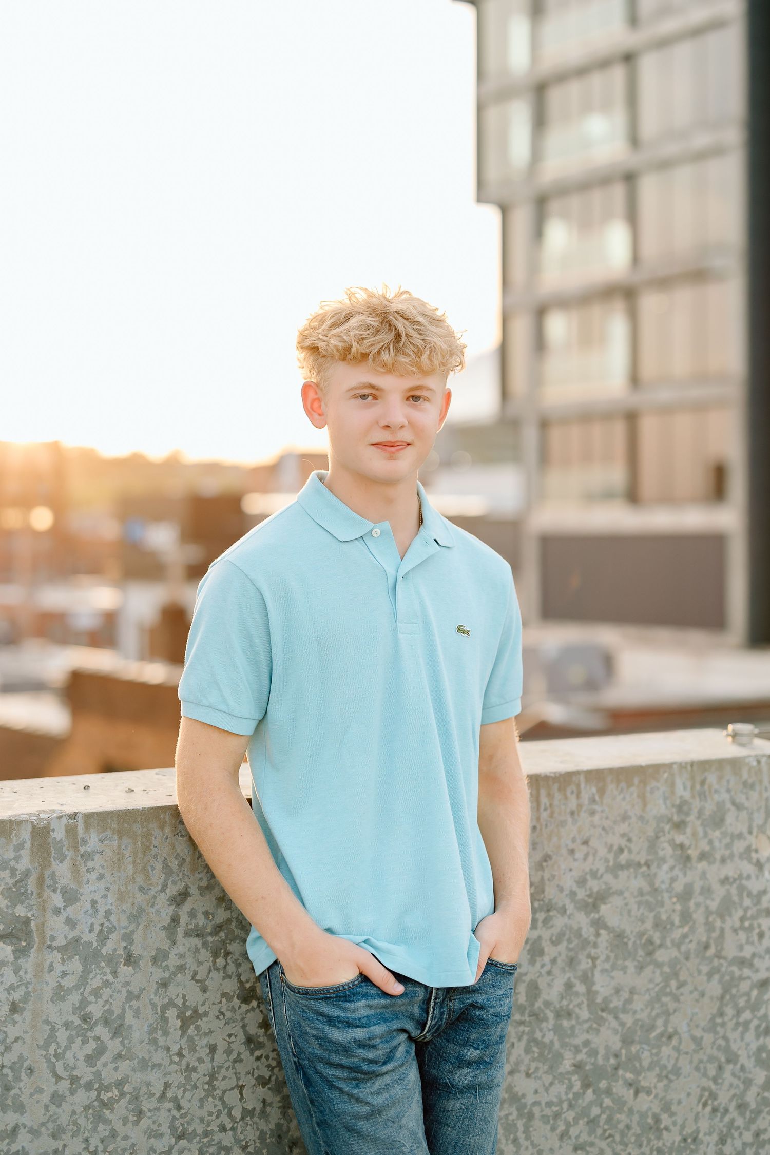 Young person in light blue polo shirt and jeans poses casually against concrete wall at sunset.