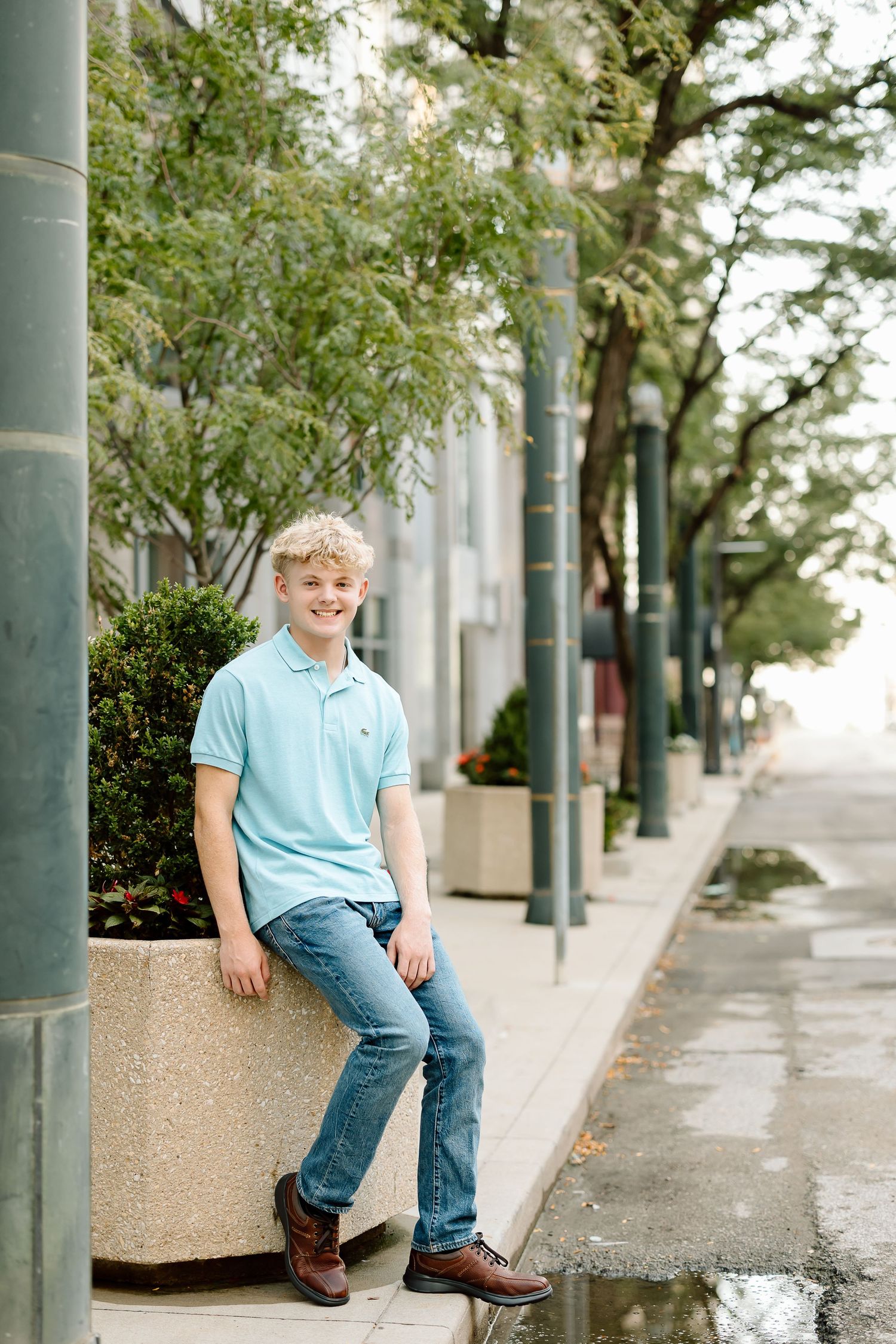 Young person in light blue shirt and jeans sits casually on concrete planter along urban sidewalk lined with trees.