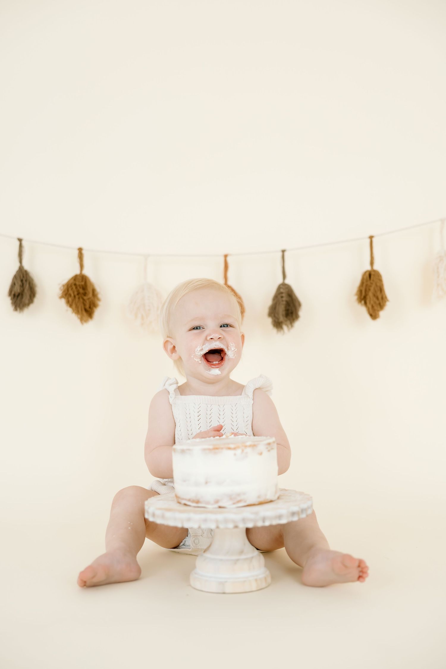 A baby wearing white sits on a white backdrop with a cake smash setup and autumn leaves hanging above.
