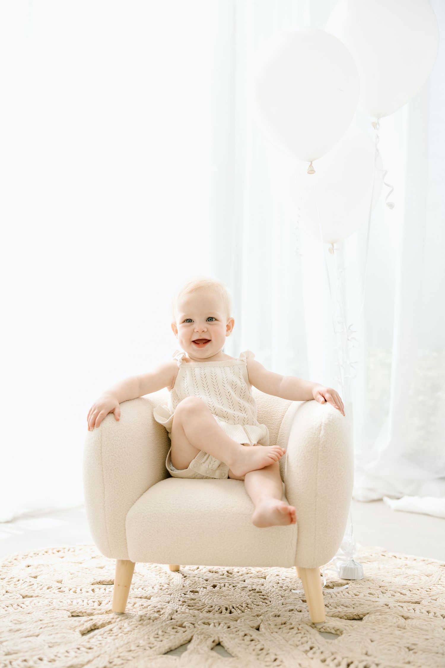 Baby sits on small white armchair against bright window backdrop, smiling and reaching out playfully.