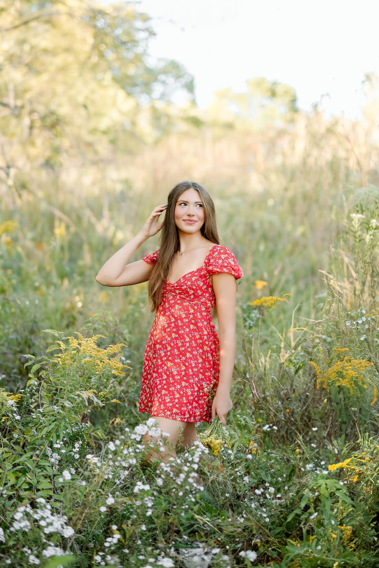 A woman in a red floral dress poses among white and yellow wildflowers in a natural meadow setting.