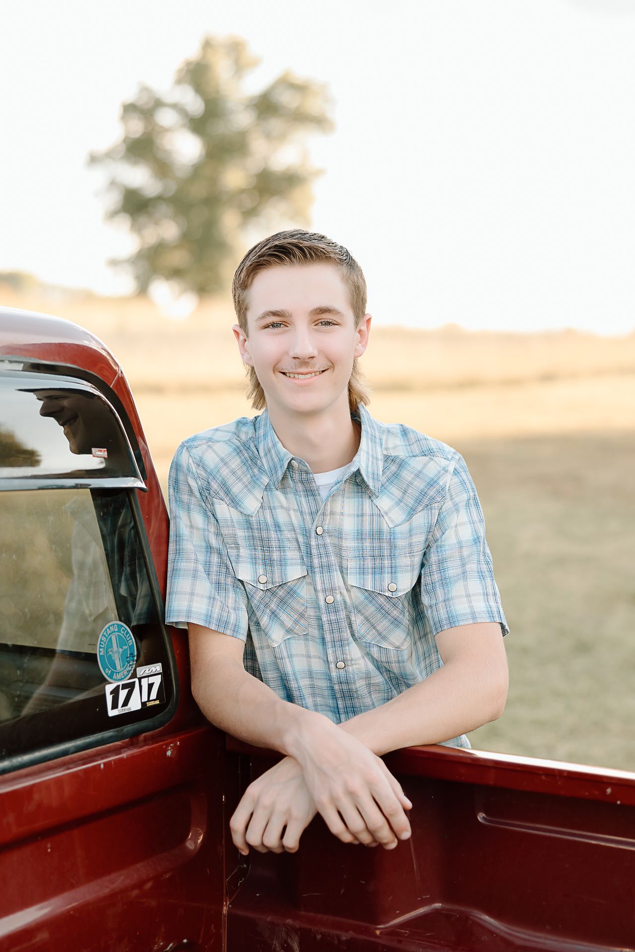 A person in a plaid shirt leans against the bed of a vintage red pickup truck in a rural setting at sunset.
