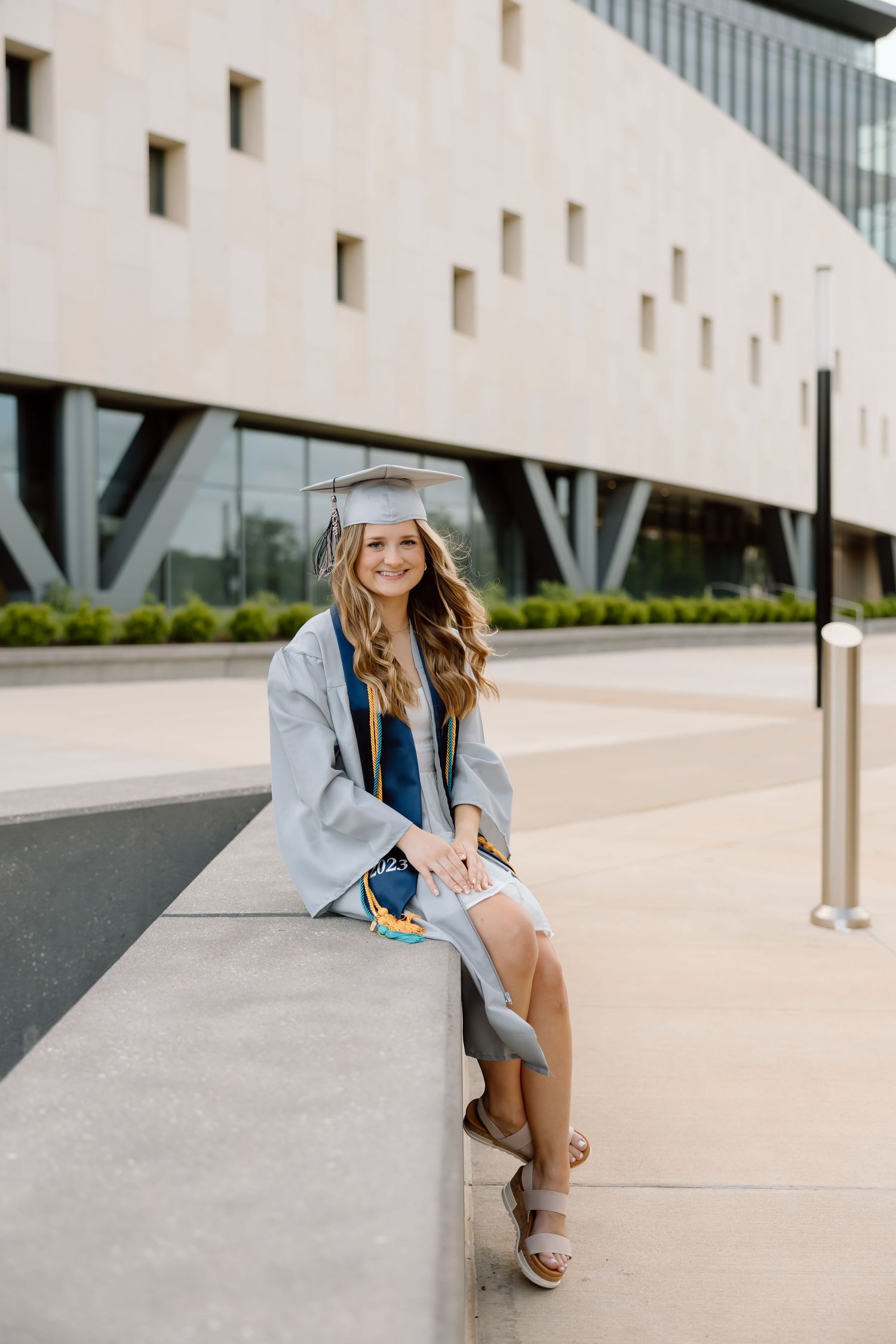 A person in a light denim jacket and brown boots sits casually on a modern concrete ledge near a white building.