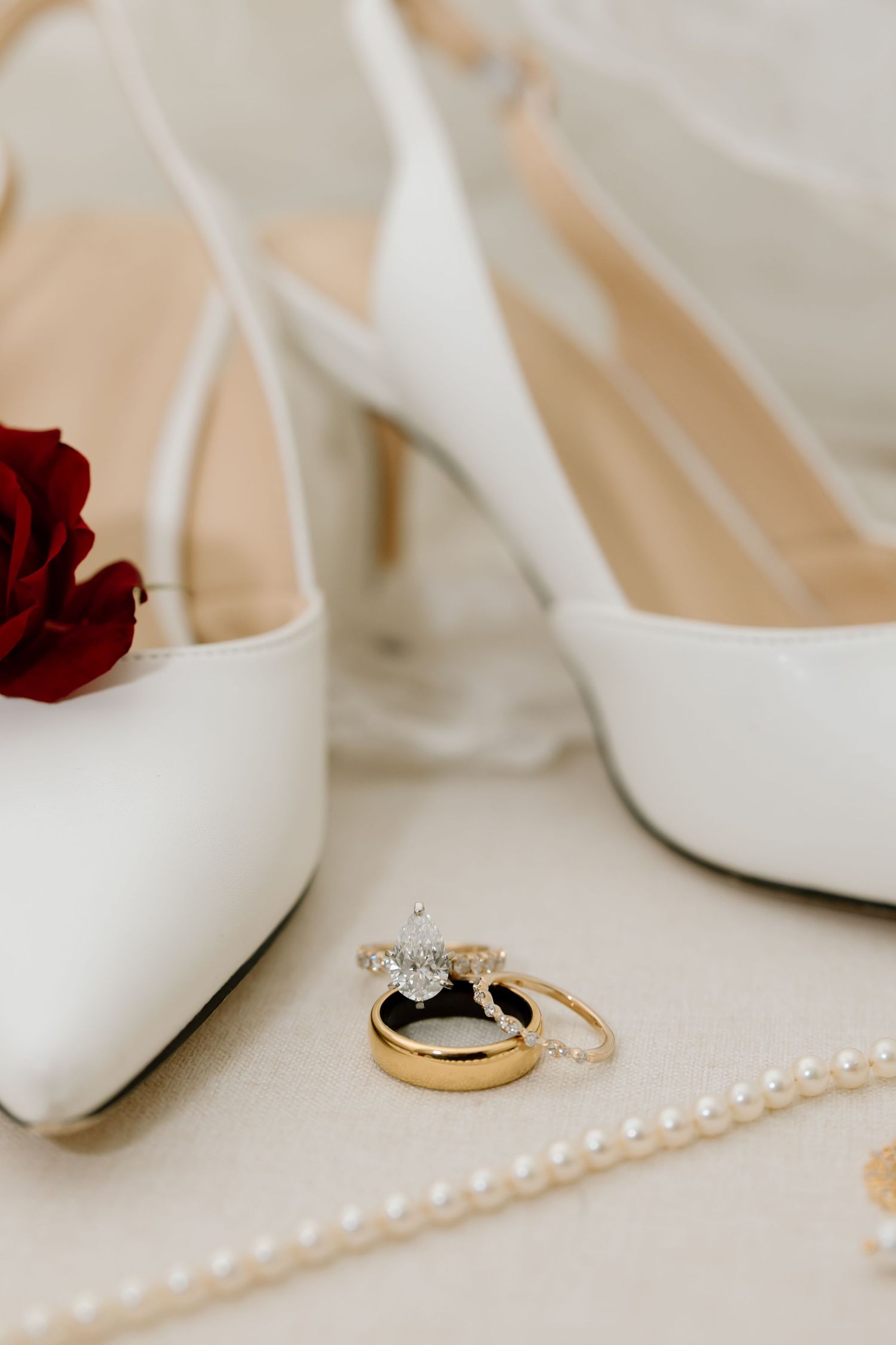 Elegant white wedding heels with gold wedding rings and a red rose on a light beige surface.