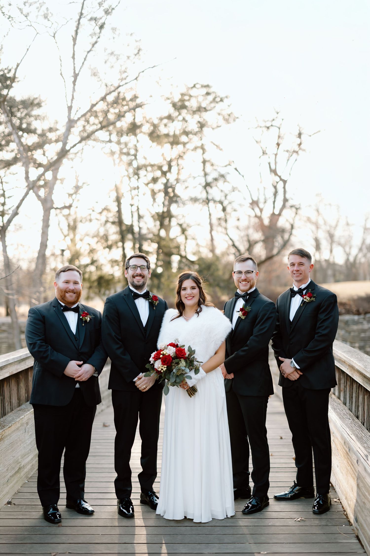 Wedding party in formal black tuxedos and white gown stand together on wooden bridge during autumn season.