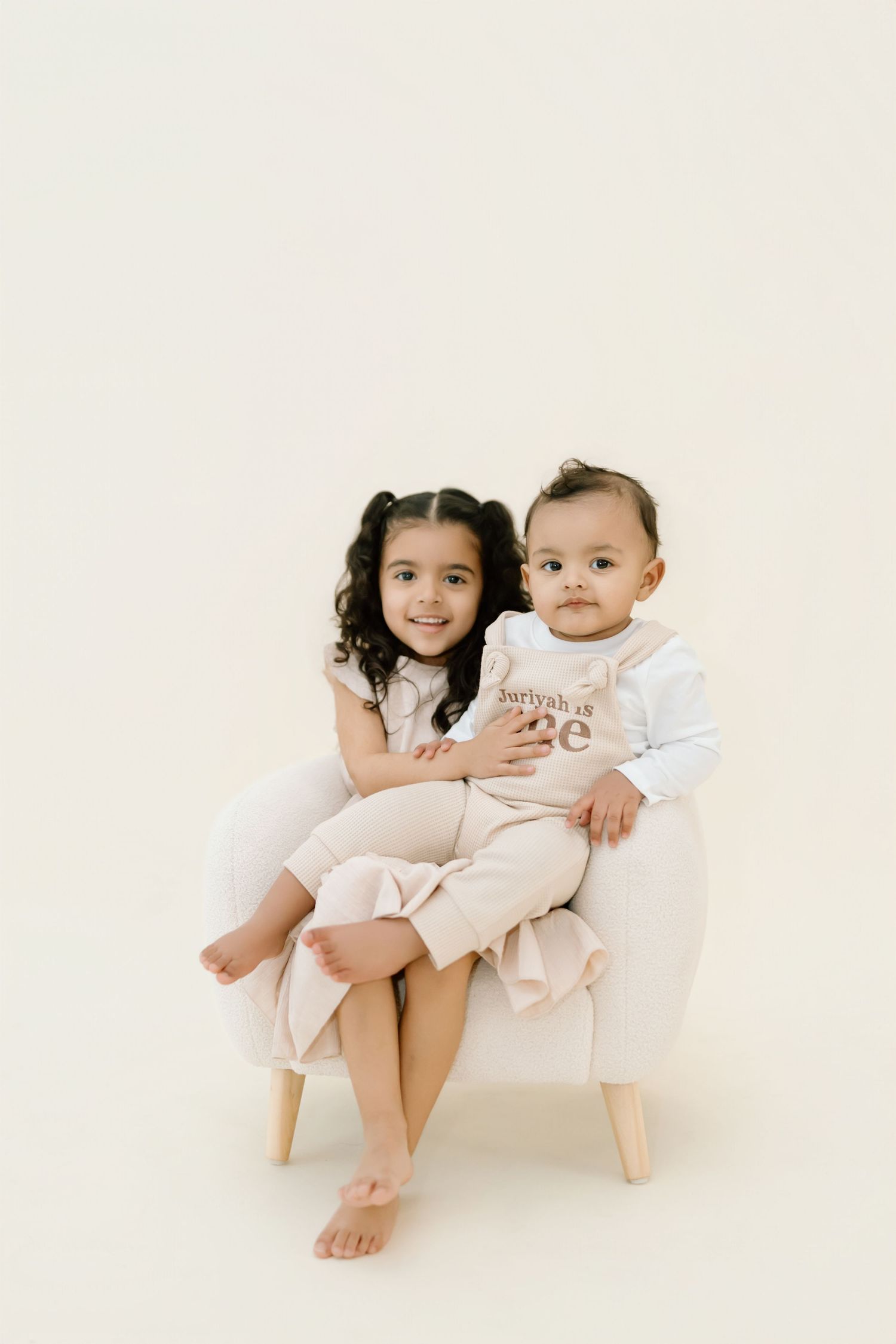 Two children share a sweet moment while sitting together on a minimalist white chair.