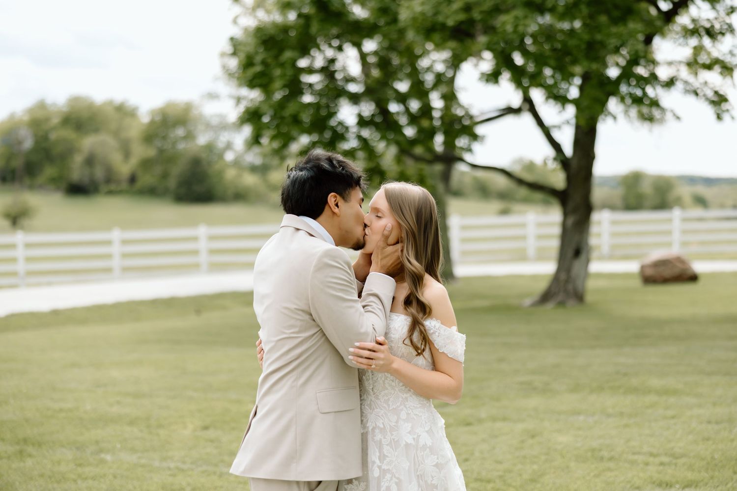 Couple sharing a romantic kiss outdoors on green lawn with white fence and trees in background.