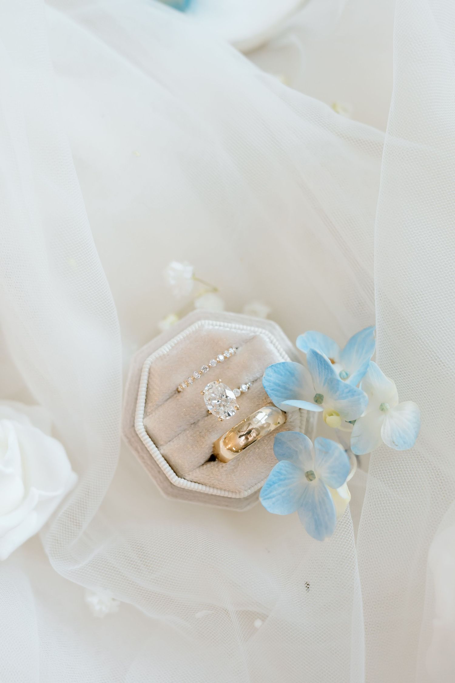 Diamond engagement ring, wedding band, and earrings in velvet box with blue hydrangeas on white veil.