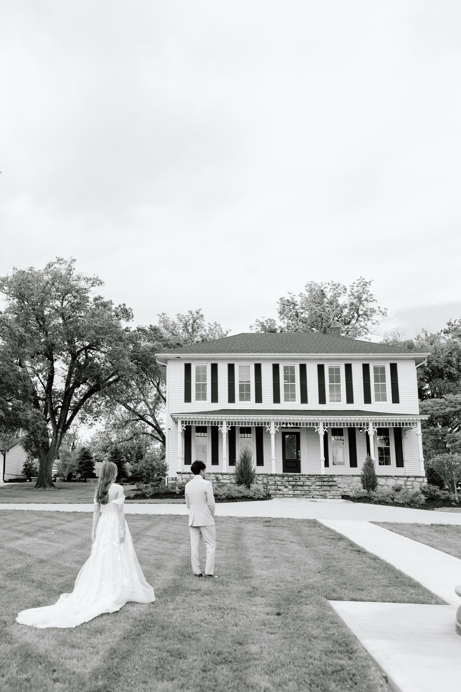 Black and white photo of a bride and groom standing before a classic two-story colonial home on their wedding day.