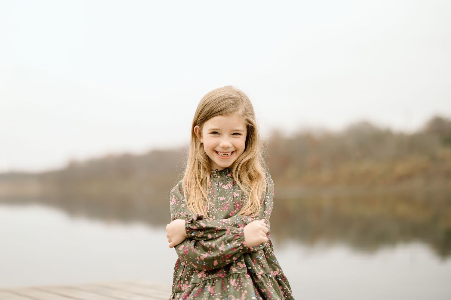 Child in a floral dress standing on a wooden dock by a lake during autumn.