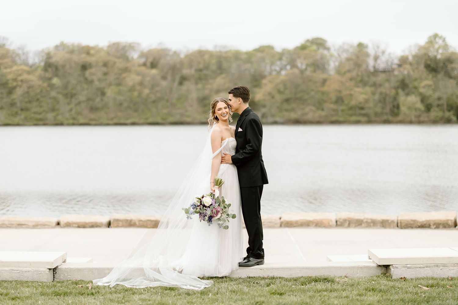 Newlyweds embrace while posing for portraits beside scenic lake.