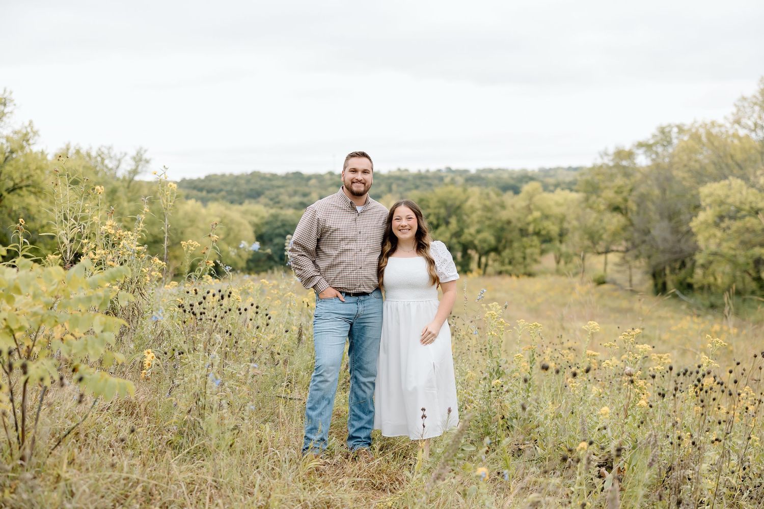 Couple stands together in a rustic field with wildflowers and rolling hills in the background.