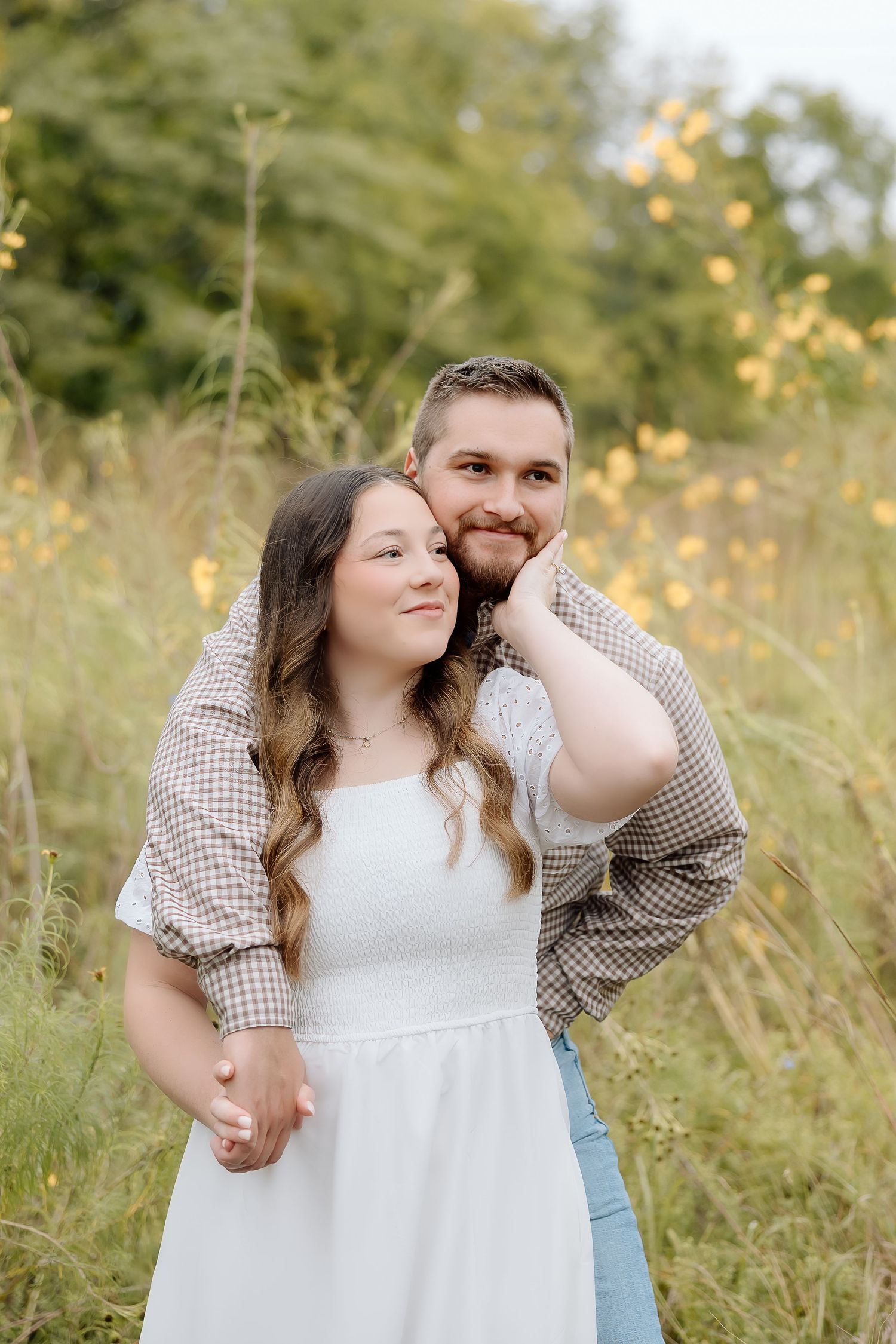 A couple poses together in casual attire among tall grass and golden wildflowers.