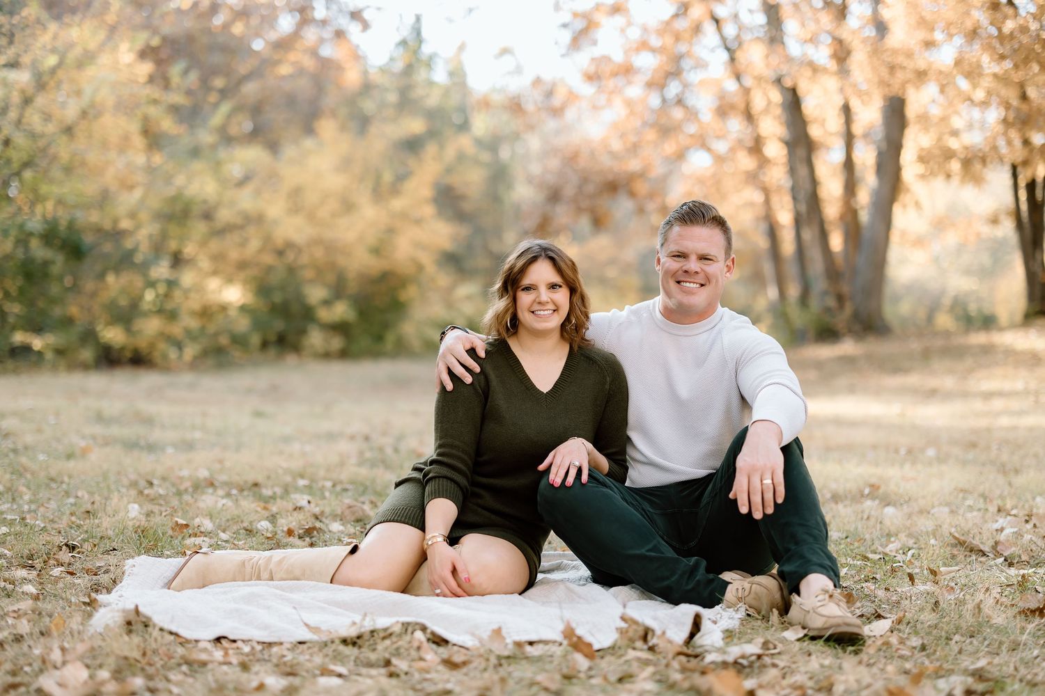 A couple sits together on a white blanket in a sunny autumn setting.