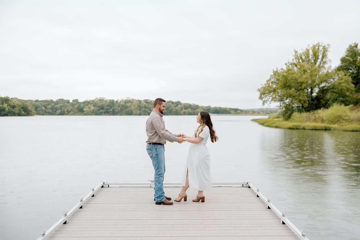 A couple holds hands while standing on a wooden dock with a lake view.