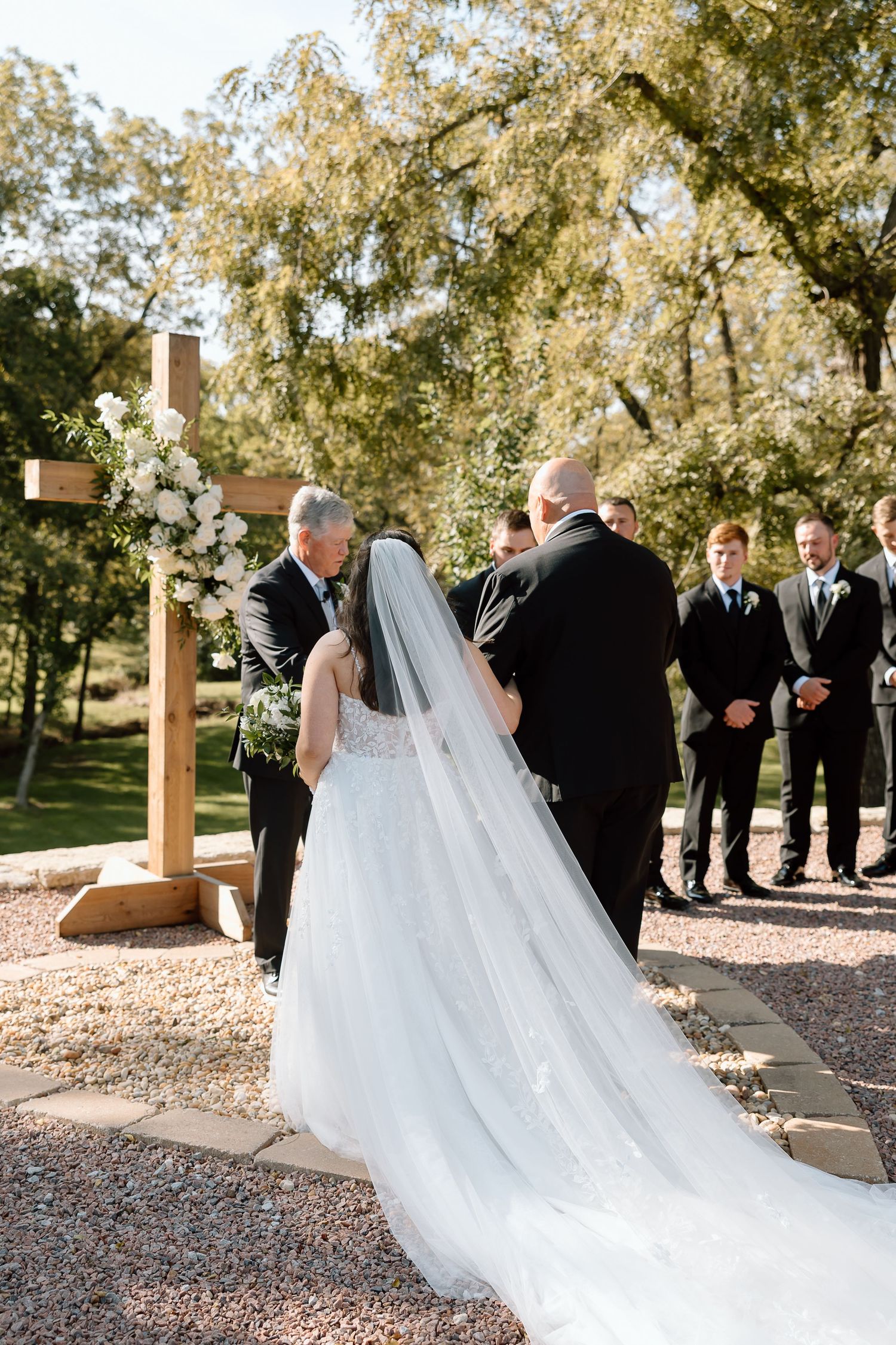 A flowing white wedding veil catches the breeze during an outdoor ceremony with wooden crosses and autumn trees.
