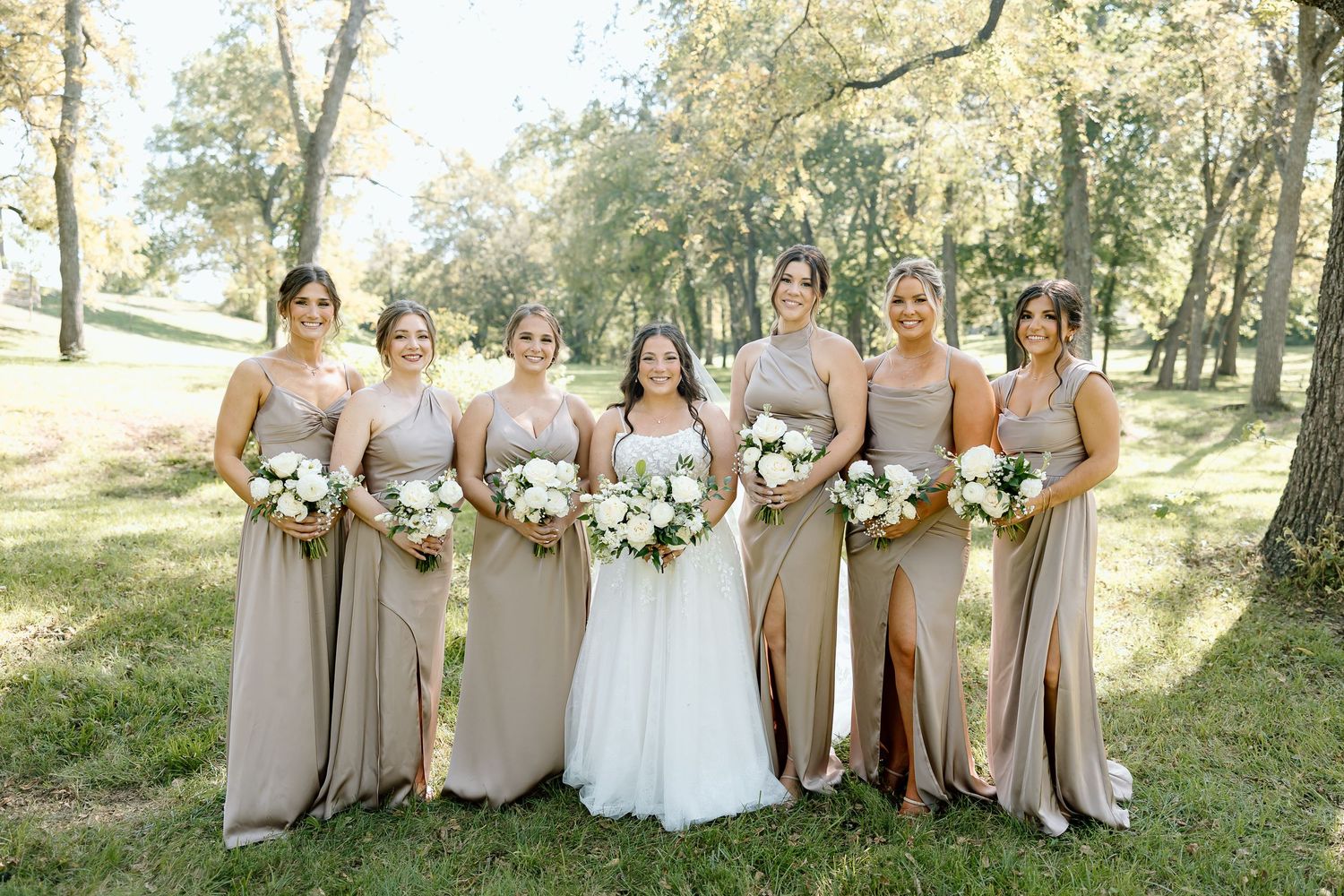 Bridal party wearing neutral taupe dresses poses together outdoors in a scenic wooded setting.