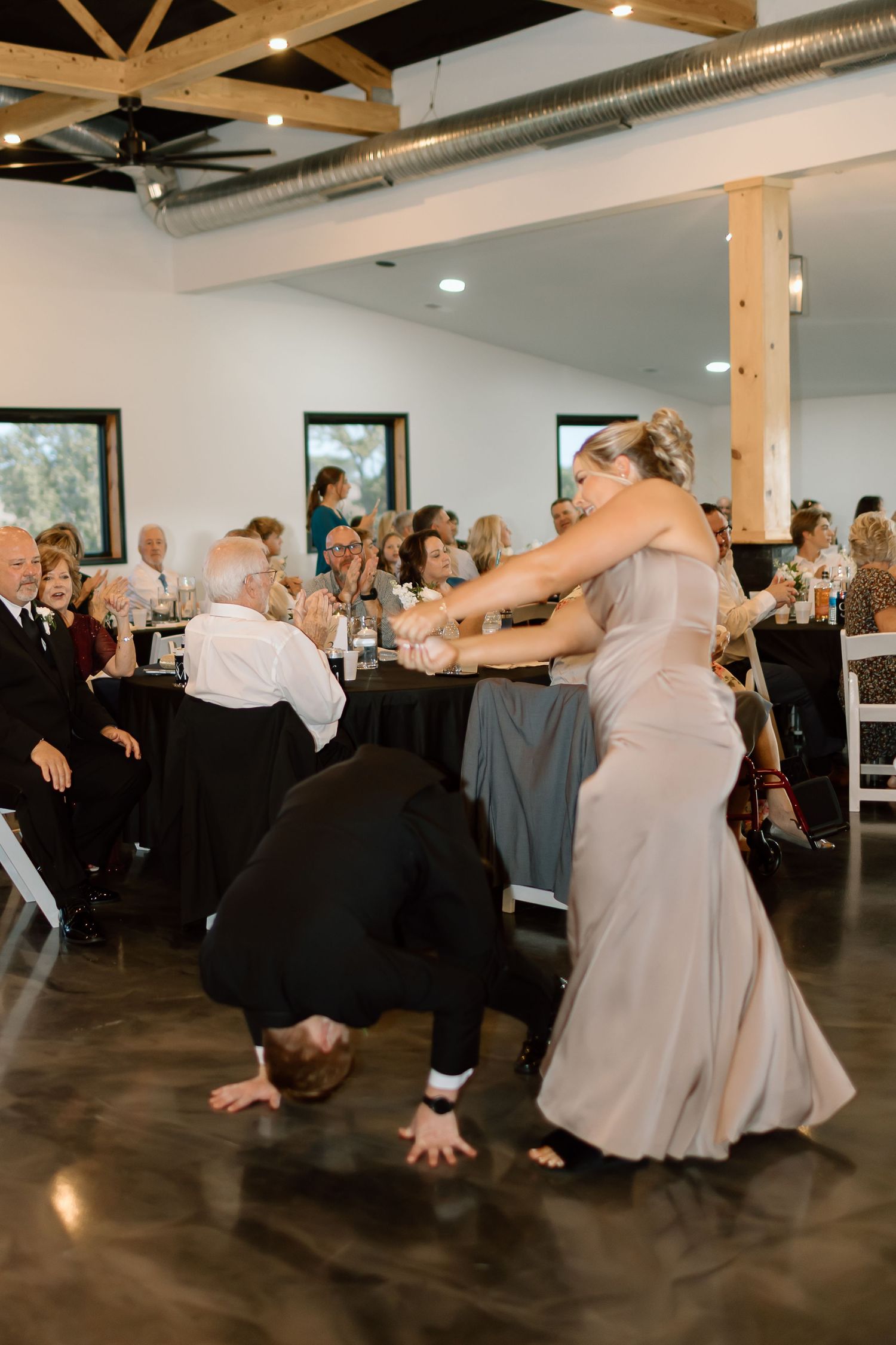 Wedding guests perform synchronized dance moves in a line at an indoor reception venue with industrial-style decor.