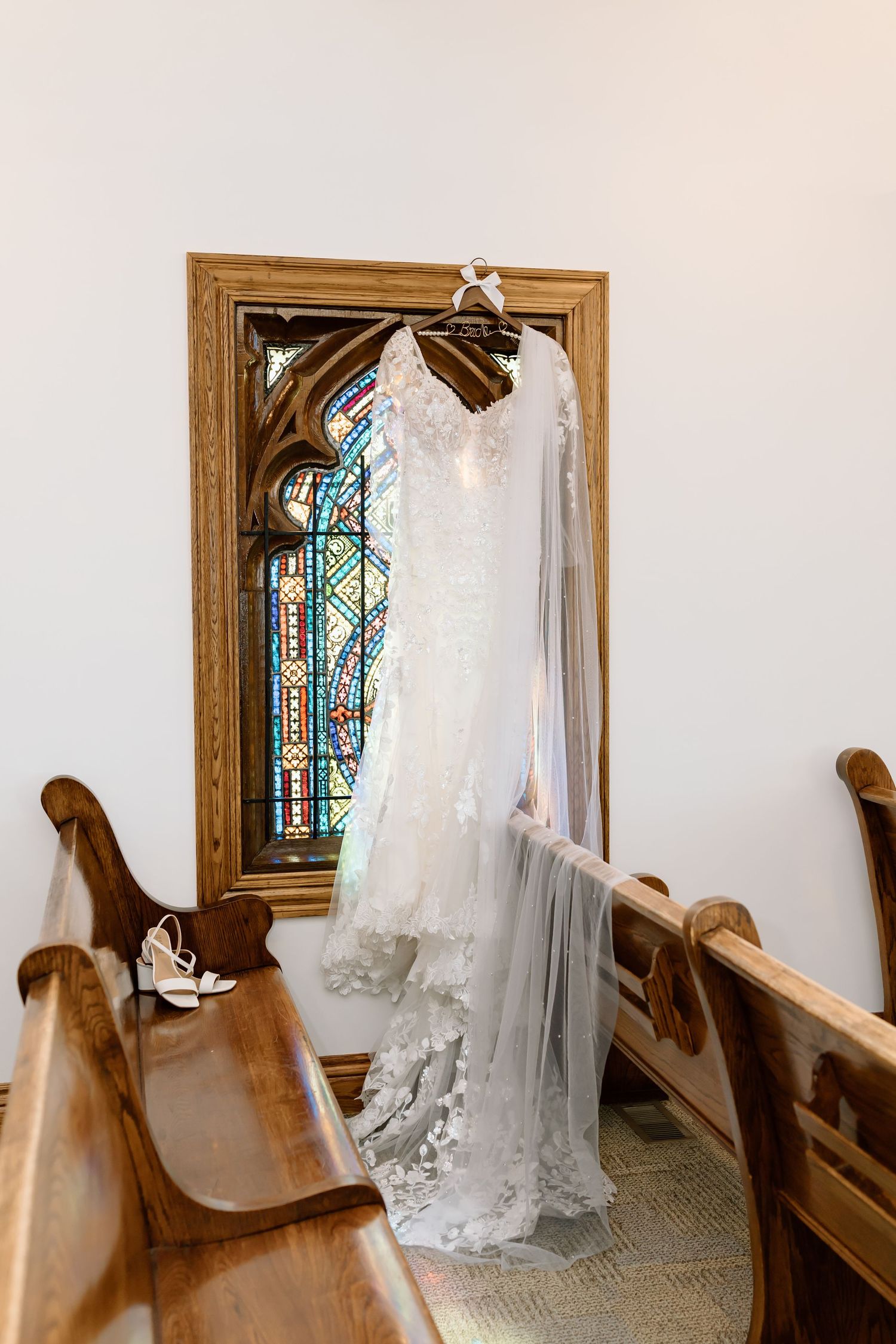 A white wedding dress hangs elegantly in a church chapel with wooden pews and stained glass windows in view.