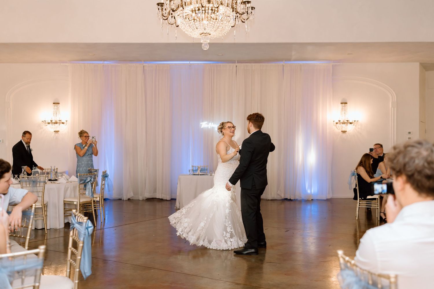 A couple shares their first dance in an elegant ballroom with white drapes and crystal wall sconces.