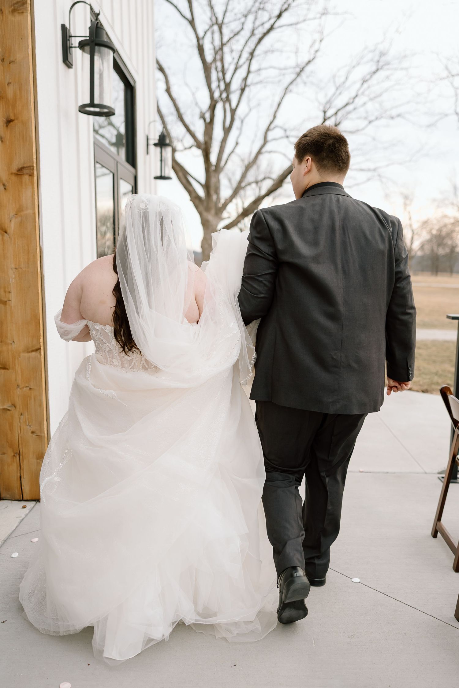 A bride in a flowing white gown and groom in dark suit walk together along a covered walkway with lanterns.