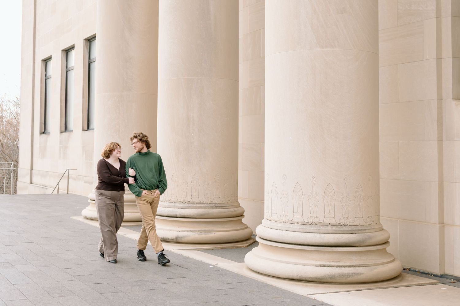 Two people sit and chat in an elegant colonnade with tall white stone columns and arched architecture.