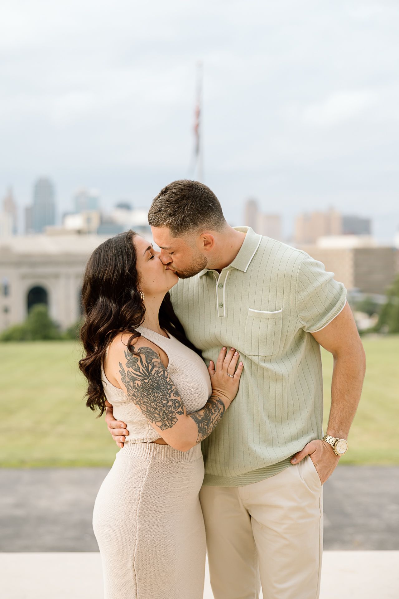 A romantic couple embraces outdoors in neutral-toned outfits against a city skyline backdrop.