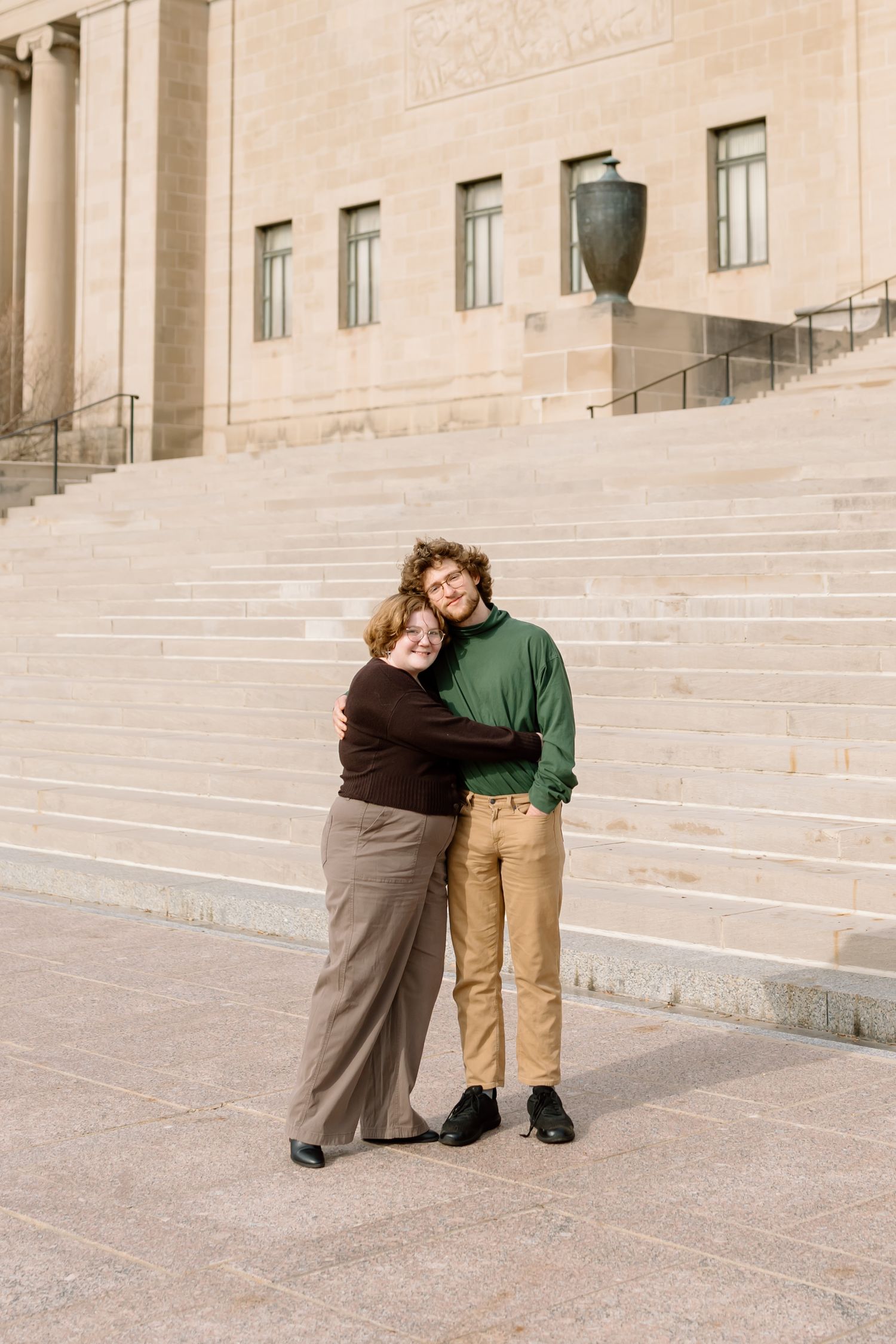 A couple embraces while standing on stone steps of a grand building with classical architecture and decorative urns.