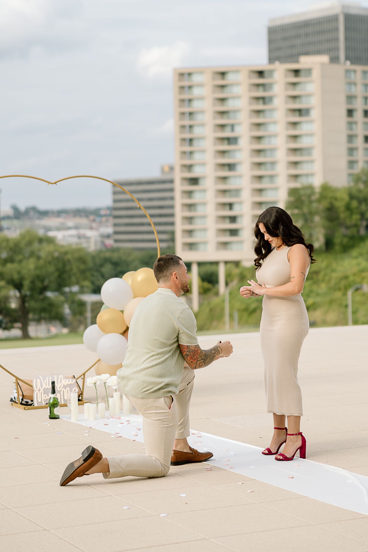 A romantic proposal scene unfolds on a rooftop with pastel balloons and a white runner against a cityscape backdrop.