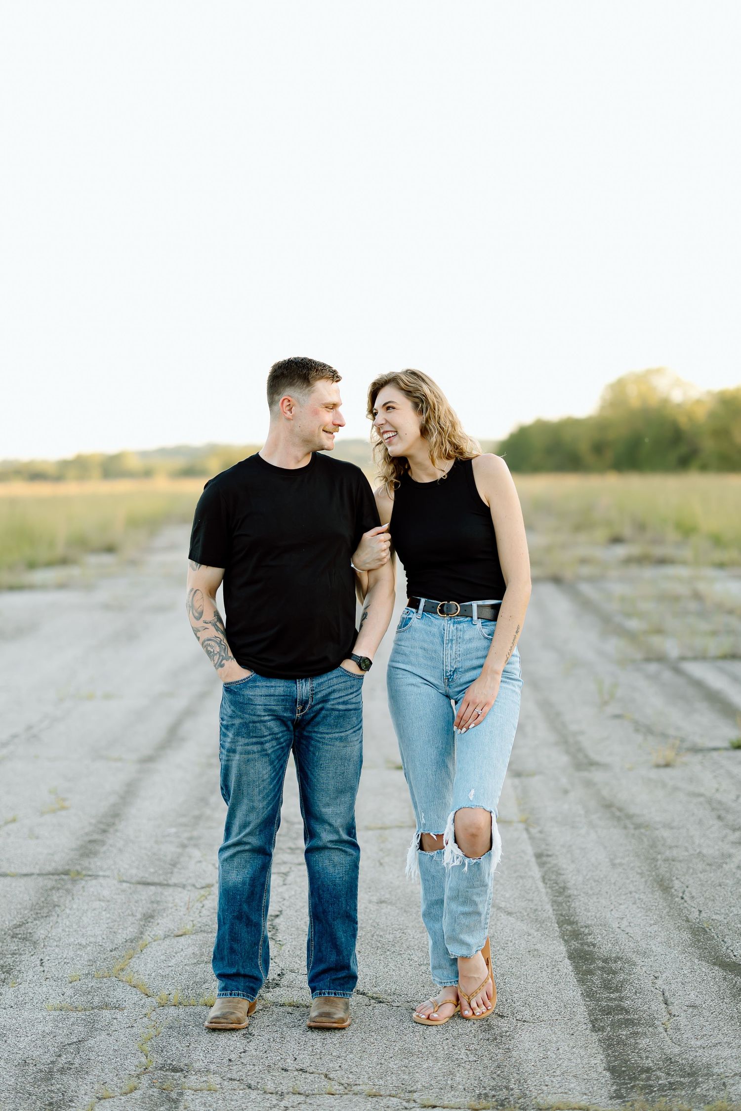 A couple in matching black shirts and blue jeans walking together on a dirt road at sunset.