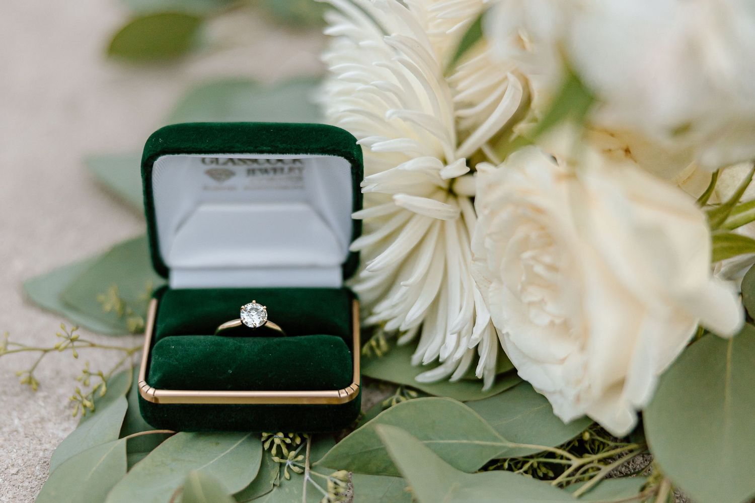 Wedding ring in green velvet box surrounded by delicate white flowers and sage green foliage on light surface.