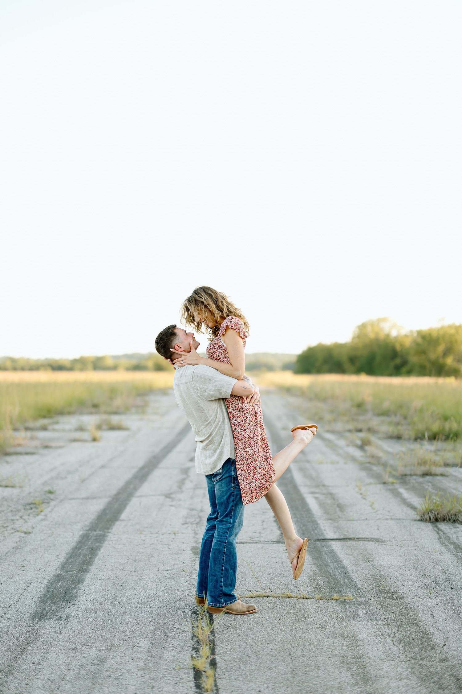A couple embraces and spins joyfully on a dirt road at sunset in a romantic countryside setting.