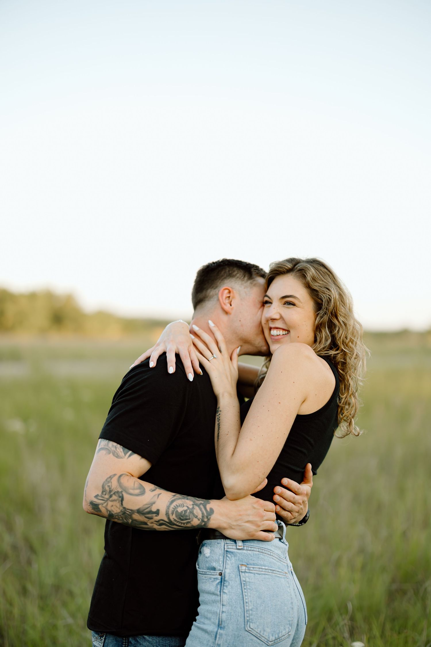 A romantic couple embraces in a sunlit meadow wearing casual attire of jeans and black shirts.