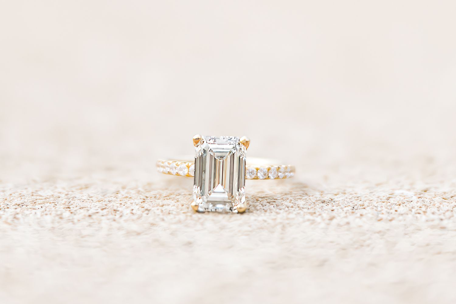 An elegant emerald-cut diamond engagement ring with delicate pave band sits on a sparkly light surface.