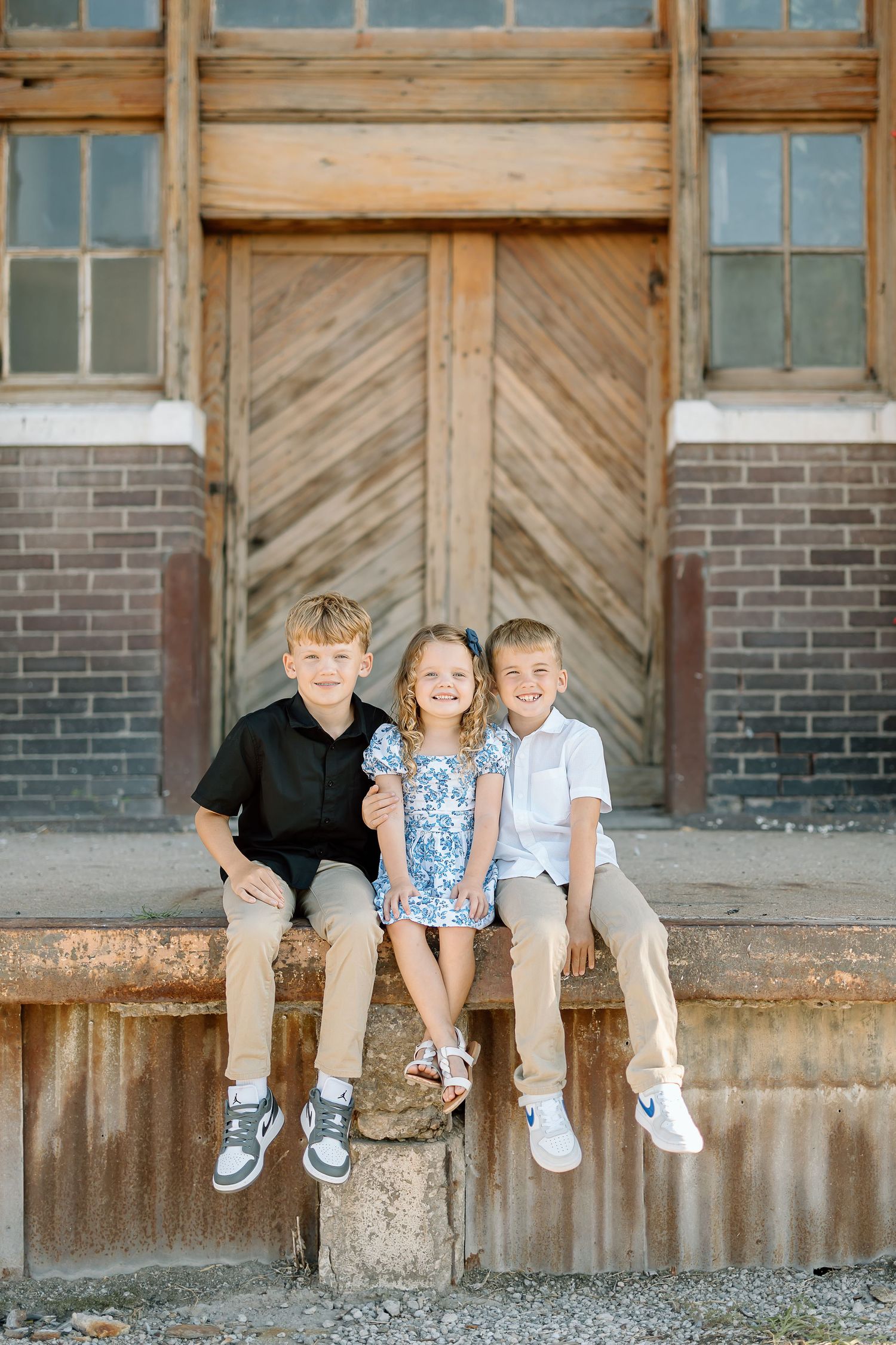 Three children sit casually on a rustic wooden ledge against a weathered barn wall with diagonal wood patterns.