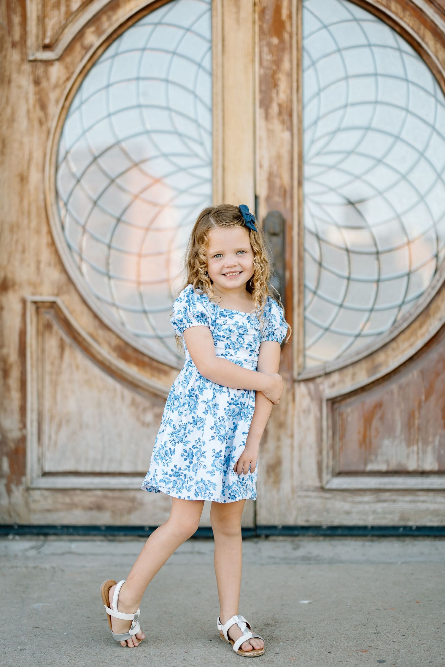 A young child in a blue and white floral dress poses playfully in front of an ornate circular door.